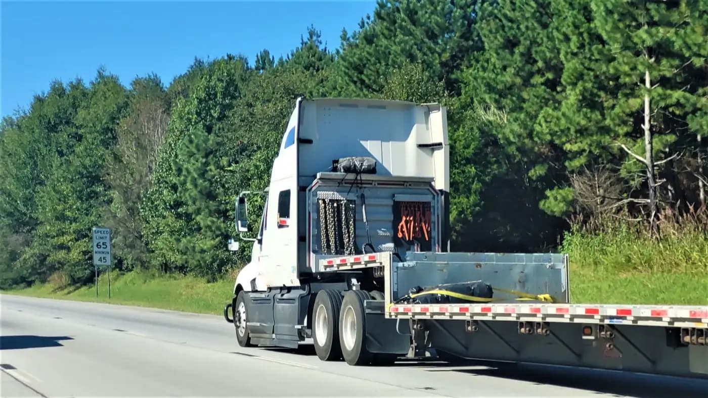 White semi-truck pulling a flatbed beside woodland